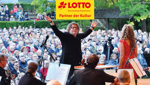 Dirigent Norddeutsche Philharmonie Marcus Bosch mit Orchester vor Publikumsmenge. Dazu Logo von LOTTO MV - Partner der Kultur.