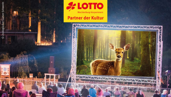 Zuschauer sitzen vor der Kulisse des Open-Air-Kino im Zoo Schwerin. Auf der Leinwand ist Bambi zu sehen.