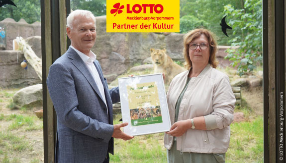 Hubert Ludwig, Geschäftsführer von LOTTO Mecklenburg-Vorpommern, und Tierparkdirektorin Katrin Töpke präsentieren vor dem Löwengehege die Patenschaftsurkunde.