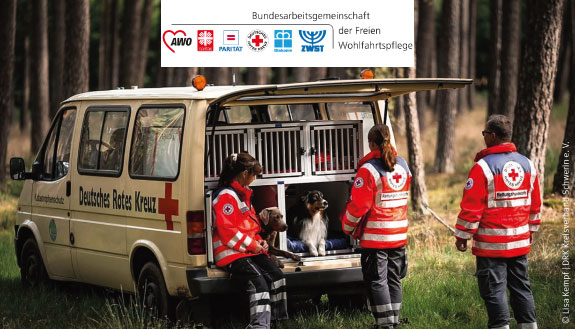Drei Mitarbeitende des DRK mit zwei Rettungshunden am Einsatzfahrzeug. Dazu Logo der Bundesarbeitsgemeinschaft der freien Wohlfahrtspflege.