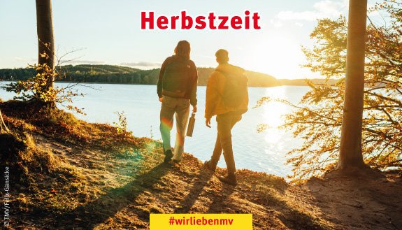 Zwei Wanderer umgeben von herbstlich gefärbten Bäumen schauen auf einen See