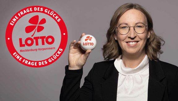 Elisabeth Aßmann, Geschäftsführerin bei LOTTO MV hält LOTTO-Kugel hoch. Dazu Logo Frage des Glücks von LOTTO MV.