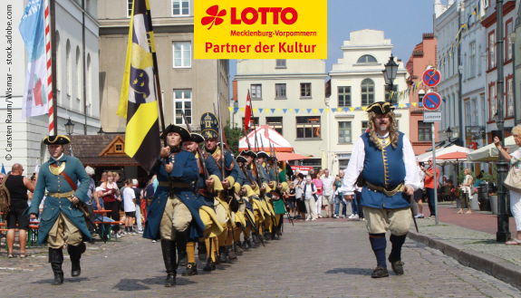 Personen in festlicher Kleidung laufen mit gro&szlig;en Fahnen eine Stra&szlig;e entlang.
