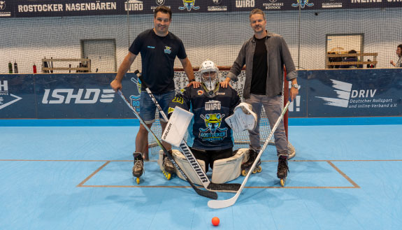 Auf dem Bild sind drei Personen in einer Halle zu sehen. Die Person in der Mitte hat eine Hockey-Ausrüstung an. Im Hintergrund befindet sich ein Tor und eine Bande.