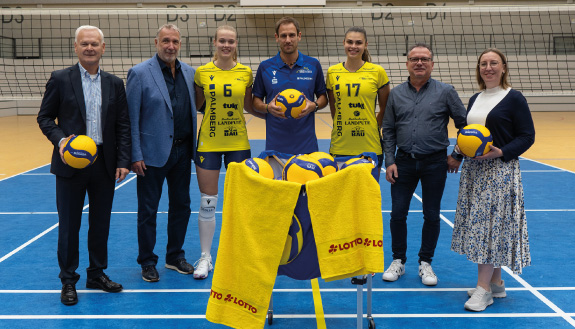 Das Bild zeigt sieben Personen. Die Personen stehen in einer Sporthalle vor einem Volleyballnetz. Drei Personen haben einen Ball in der Hand. Vor den sieben Personen steht ein blauer Korb mit Bällen und zwei Handtüchern.